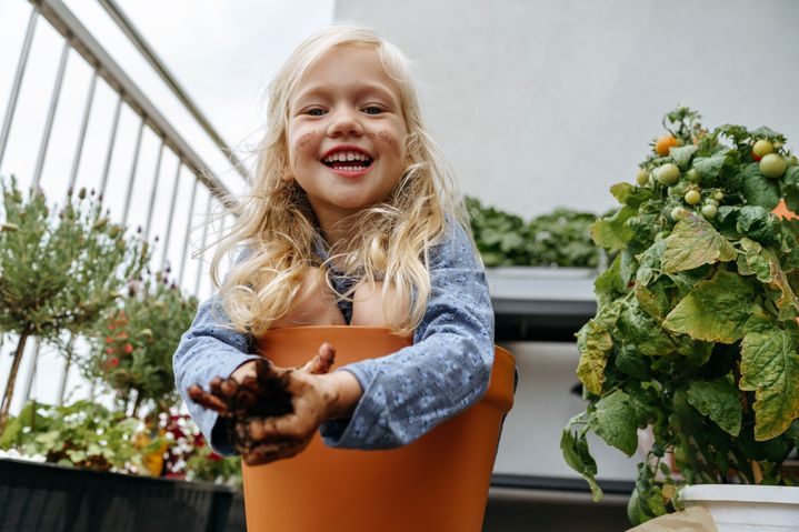 Cheerful girl with dirty hands sitting in pot near plants at balcony