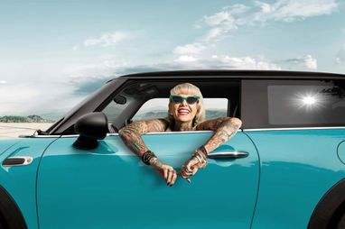 Tätowierte Frau mit Sonnenbrille schaut breit lächelnd aus einem türkisblauen Mini Cooper.