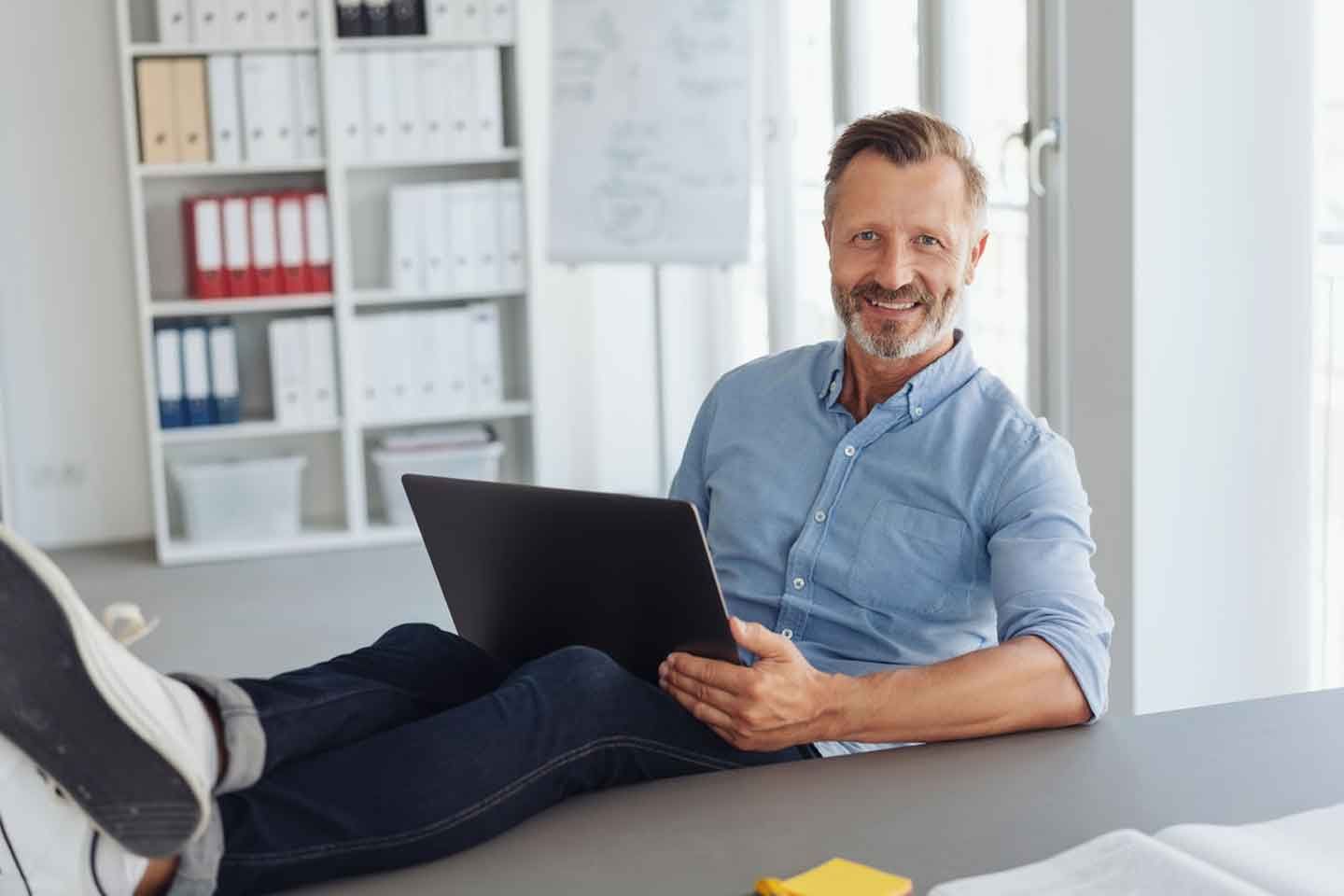 Ein lächelnder Mann in lässiger Haltung sitzt mit hochgelegten Beinen am Schreibtisch und arbeitet entspannt am Laptop.