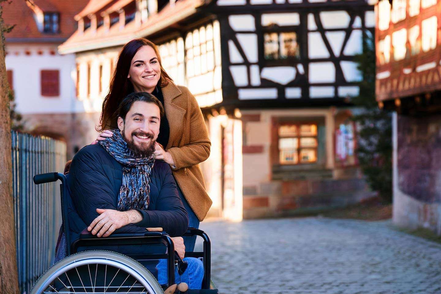 Ein Mann im Rollstuhl und eine Frau stehen lächelnd vor einem historischen Fachwerkhaus in einer Altstadtgasse.