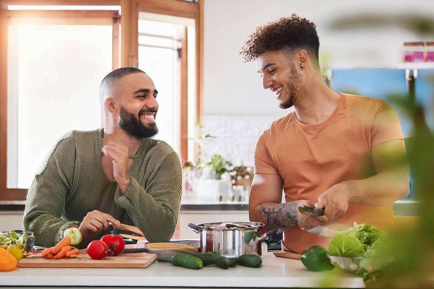 Zwei Männer stehen lachend in einer Küche und bereiten gemeinsam frisches Gemüse zu.