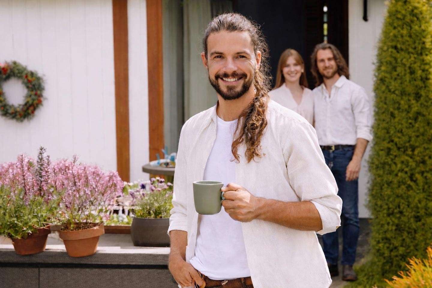 Ein Mann mit langem Haar hält lächelnd eine Tasse im Garten