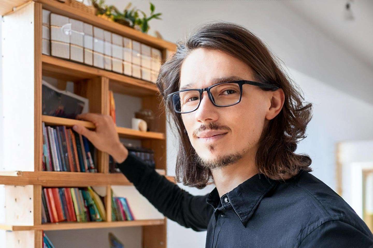 Ein Mann mit Brille steht vor einem Bücherregal und greift nach einem Buch.