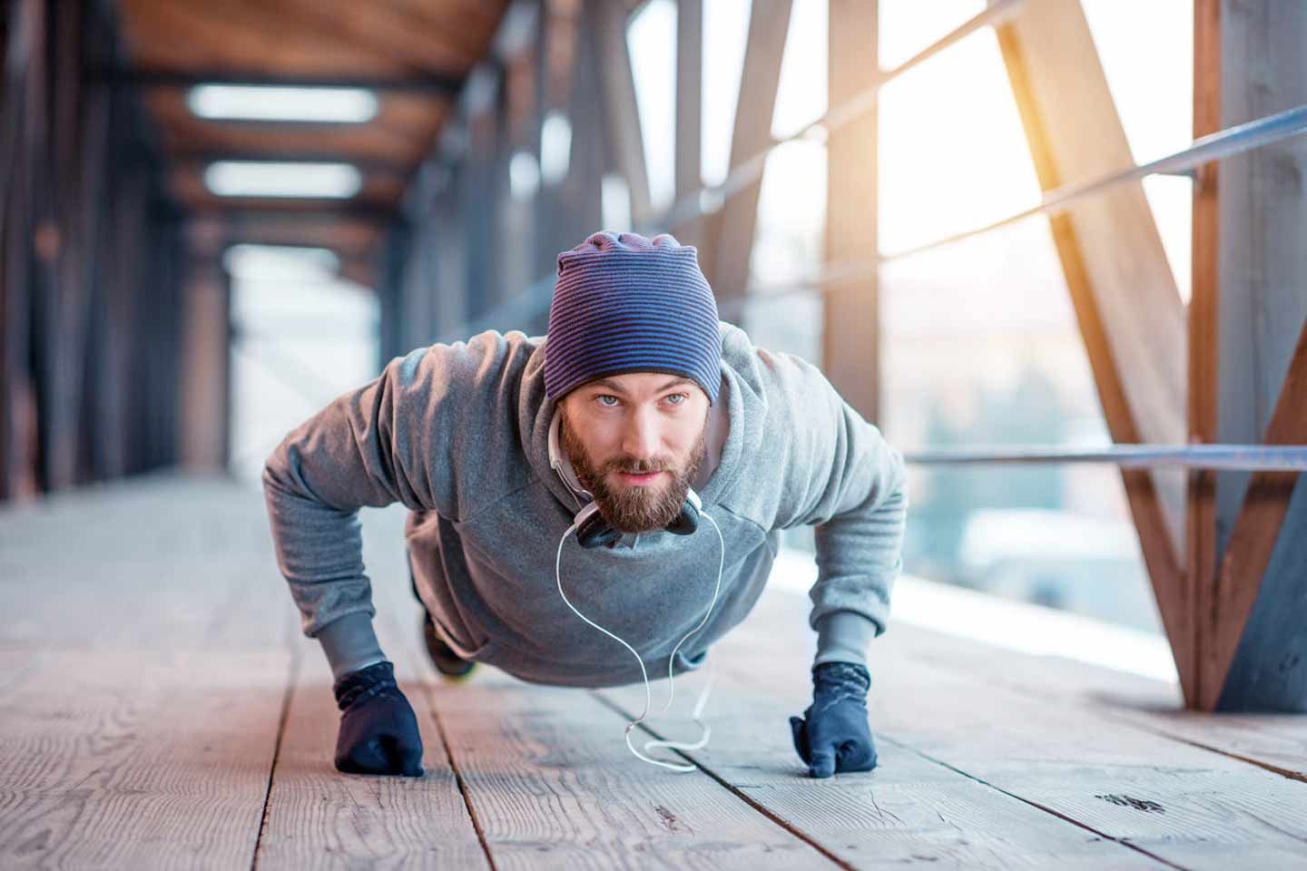 Ein sportlicher Mann macht im Winteroutfit Liegestütze auf einer Holzbrücke bei Sonnenlicht..