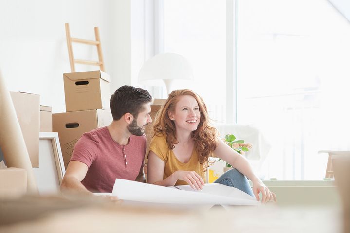 Ein junges Paar sitzt zwischen Umzugskartons in einer hellen Wohnung und schaut gemeinsam auf einen Plan oder eine Zeichnung.