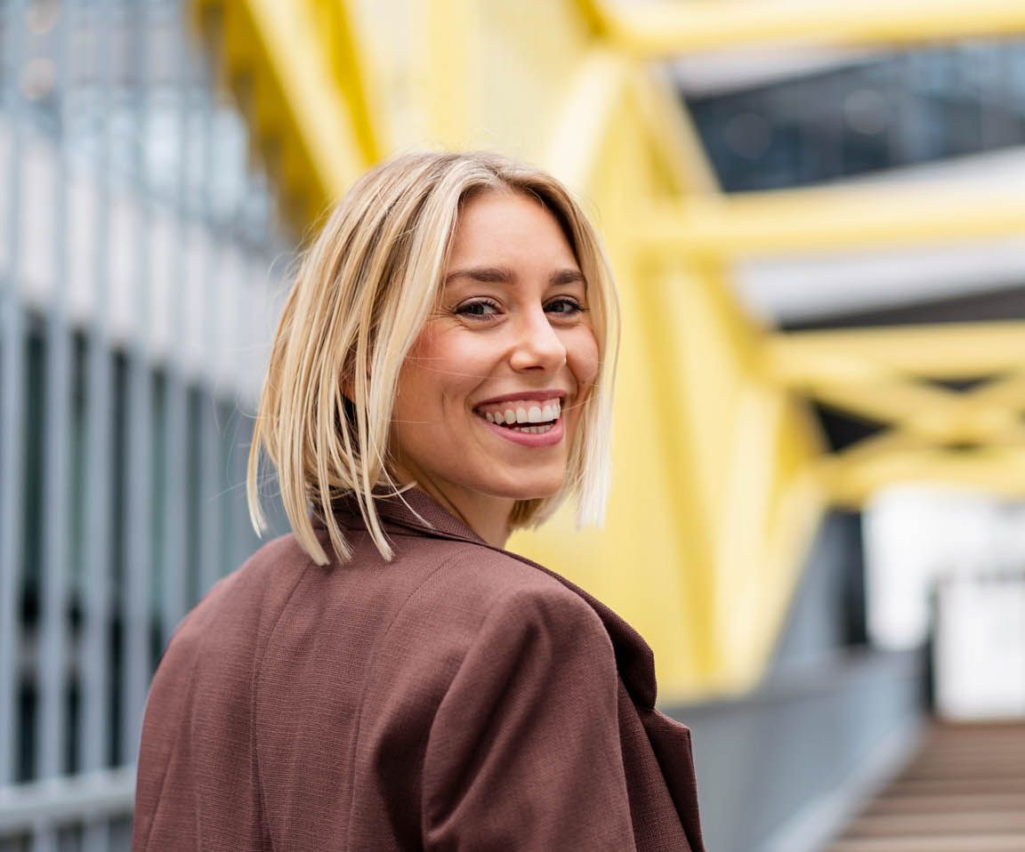 Eine Blondine mit Bob lächelt über die Schulter während sie über eine Brücke geht. 