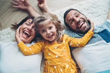 Eltern liegen auf dem Teppichboden und lachen mit der kleinen Tochter in die Kamera. 