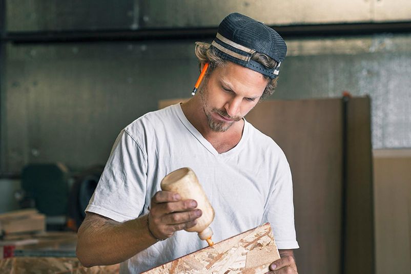 Ein Schreiner trägt konzentriert Leim auf ein Holzbrett in seiner Werkstatt auf.