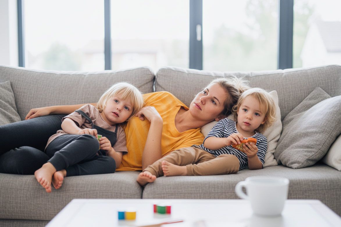 Mutter liegt entspannt auf einem grauen Sofa, zwei kleine Kinder kuscheln links und rechts an ihr.