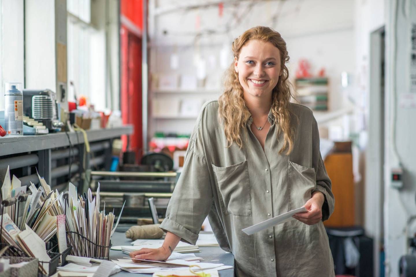 Eine lächelnde Frau steht in einem kreativen Arbeitsraum zwischen Papierstapeln und Werkzeugen und hält ein Blatt in der Hand.