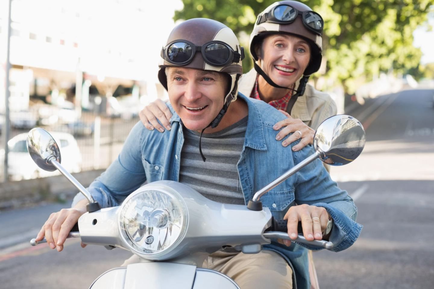 Älteres Paar mit Helmen fährt gemeinsam auf einen Motorroller und genießt die Fahrt bei sonnigen Wetter.