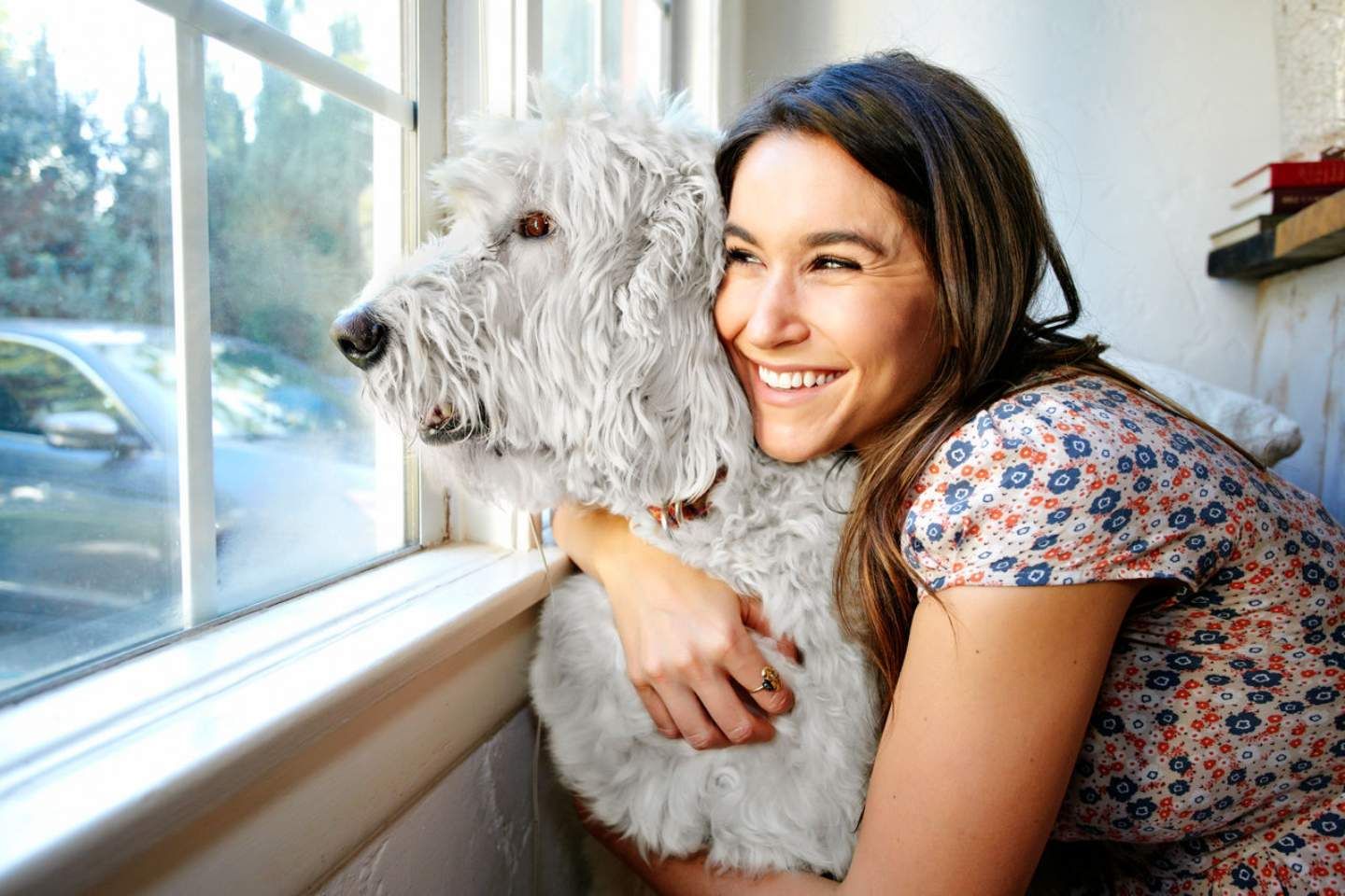 Frau umarmt ihren weißen Hund vor einem Fenster