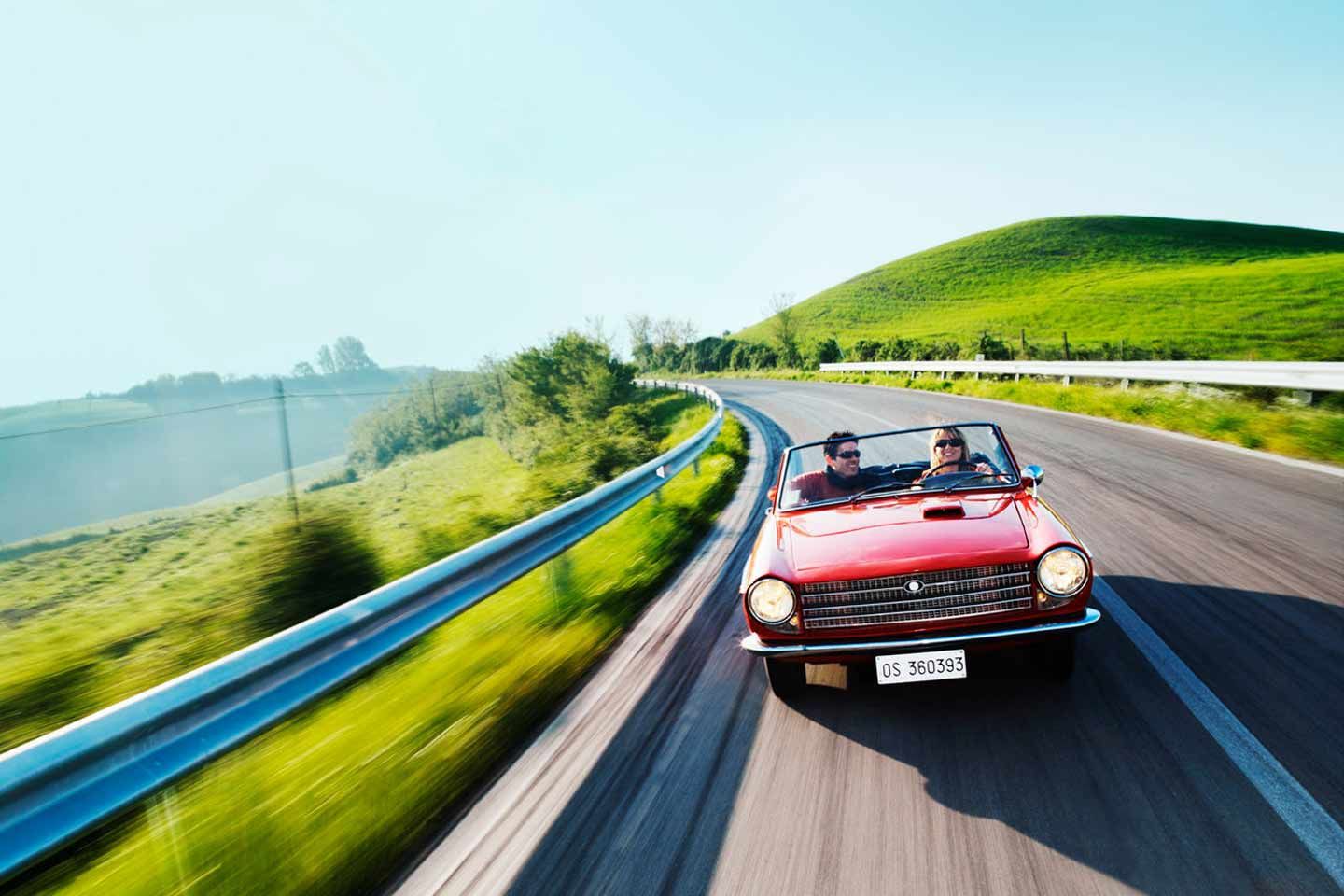 Roter Oldtimer mit zwei Personen fährt durch italienische Landschaft.