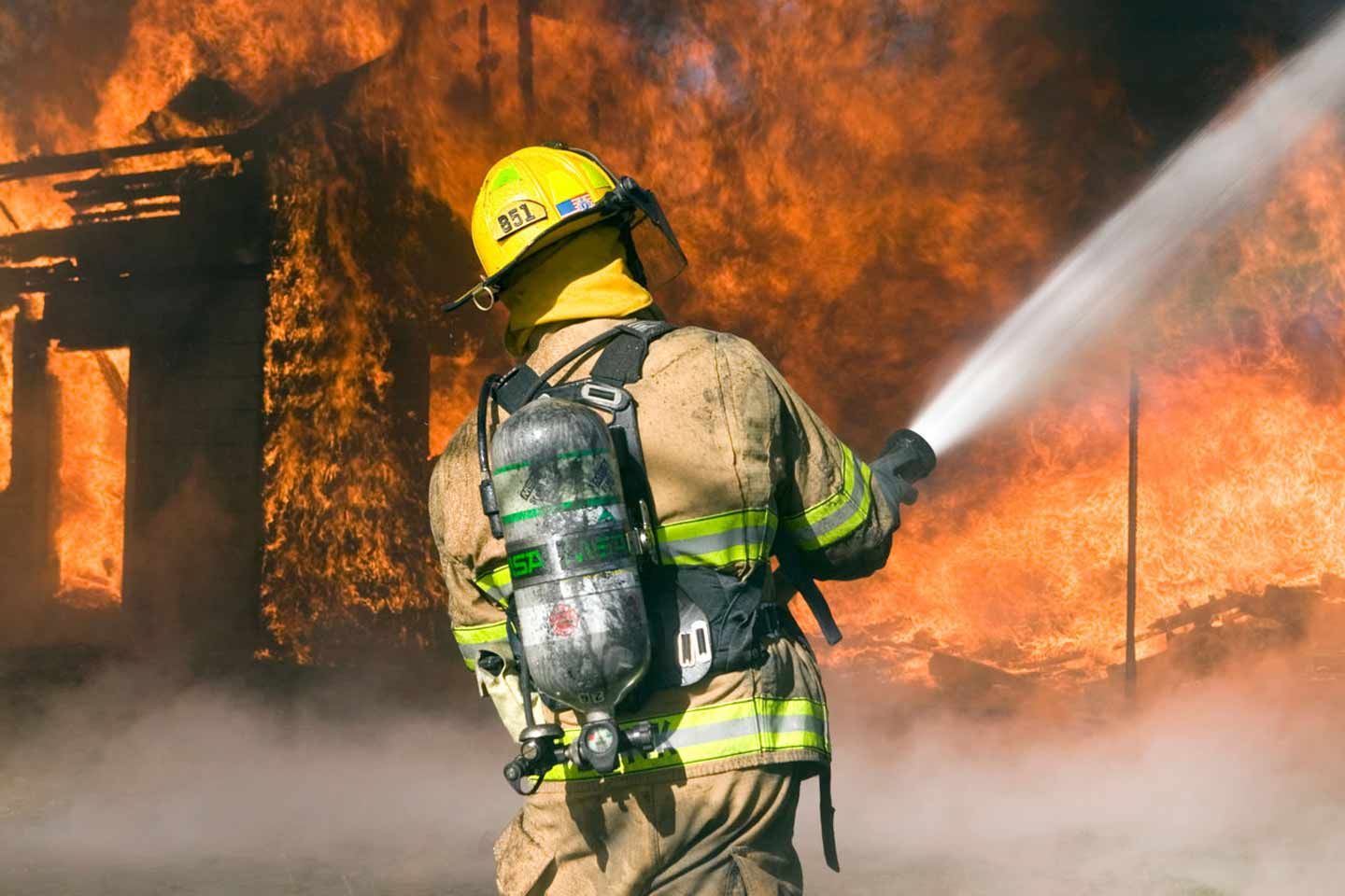 Ein Feuerwehrmann löscht ein brennendes Gebäude mit einem Wasserstrahl - Rauch und Flammen im Hintergrund.