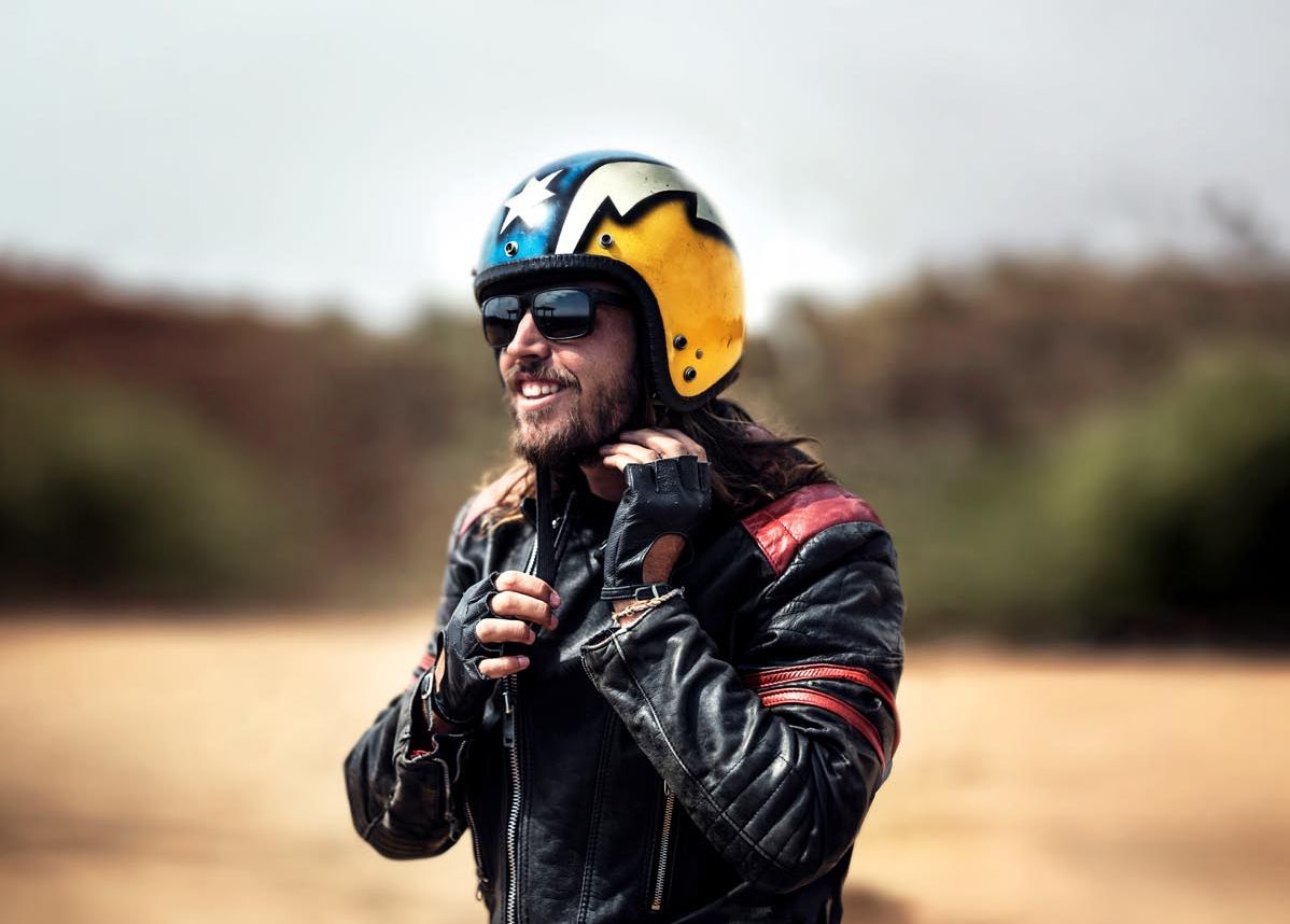 Bärtiger Mann mit einer schwarzer Lederjacke und Sonnenbrille, der seinen  bunten offenen Sturzhelm einstellt.