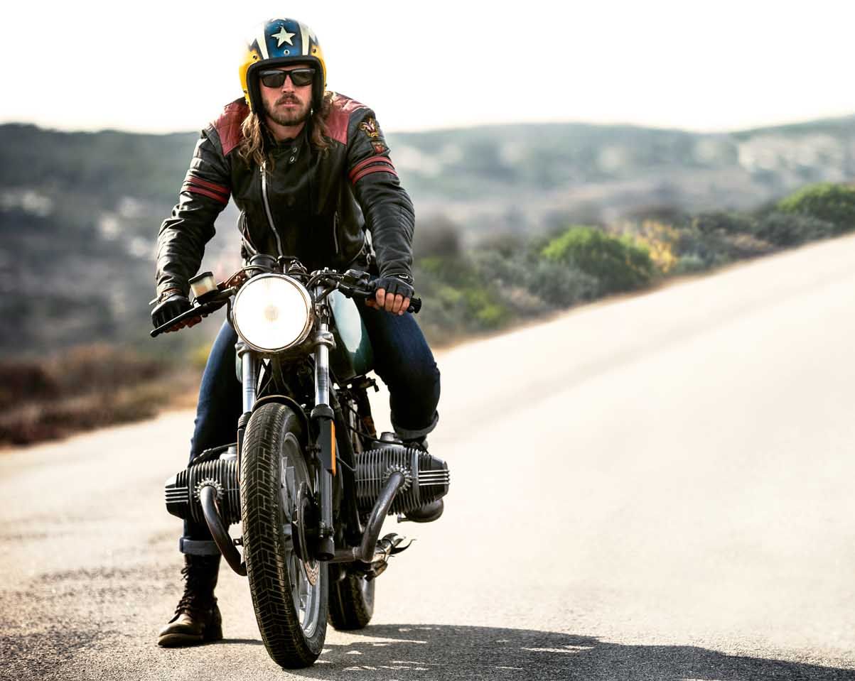 Ein Mann mit offenen Sturzhelm und Schutzbrille sitzt auf Cafe-Racer-Motorrad auf einer Landstraße.