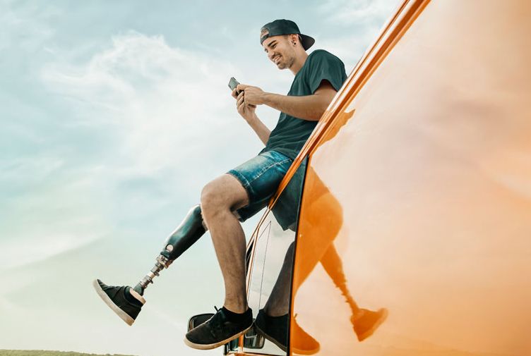 Mann mit Prothese sitzt am orangefarbenen Van vor Landschaft und Himmel.