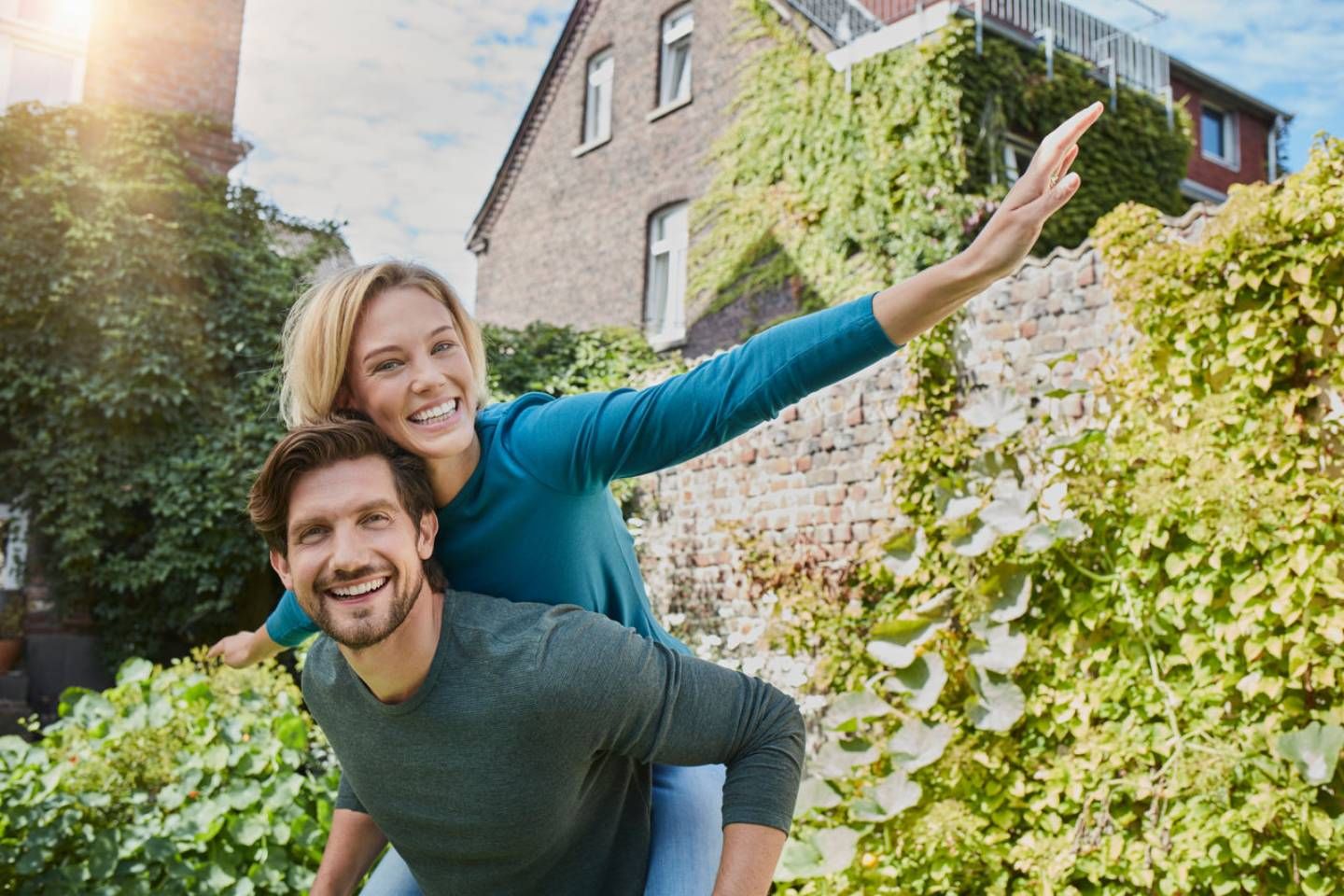 Paar im Garten, Frau auf dem Rücken des Mannes, beide lachen in die Kamera.