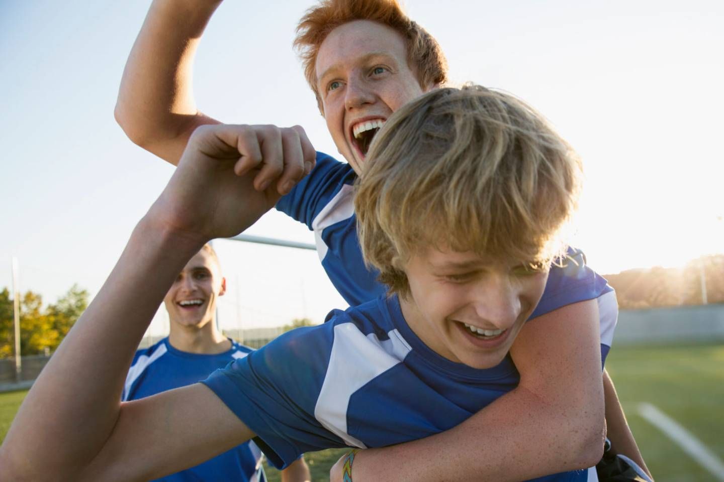 Zwei junge Männer in Sportkleidung feiern ausgelassen auf einem Fußballfeld, einer trägt den anderen huckepack.