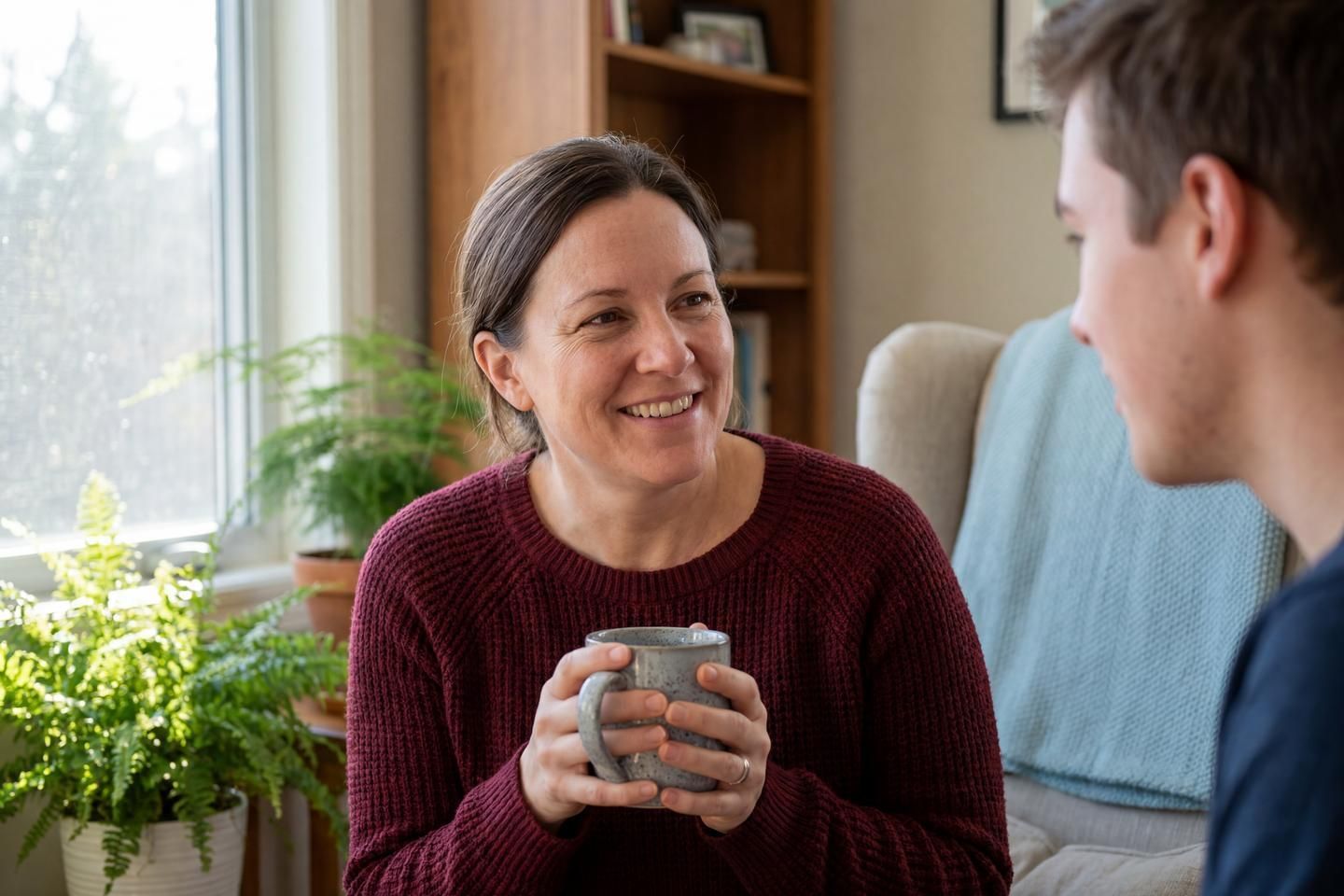 Eine Person sitzt in einem hellen Wohnzimmer, hält eine Tasse in den Händen und spricht mit einer zweiten Person im Vordergrund.