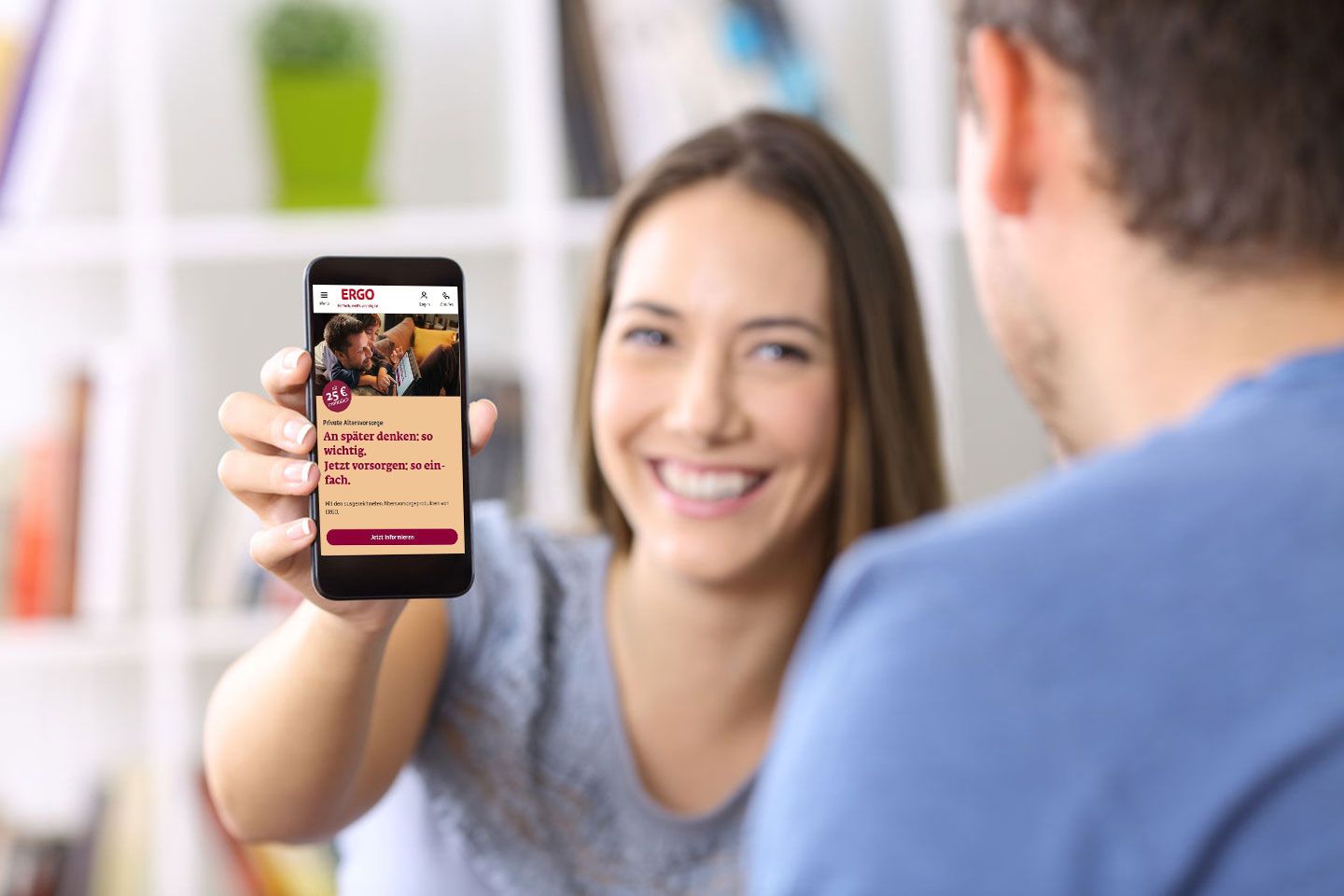 Eine Frau lächelt in Richtung Kamera und hält ein Smartphone hoch, auf dem die ERGO-Webseite geöffnet ist.
