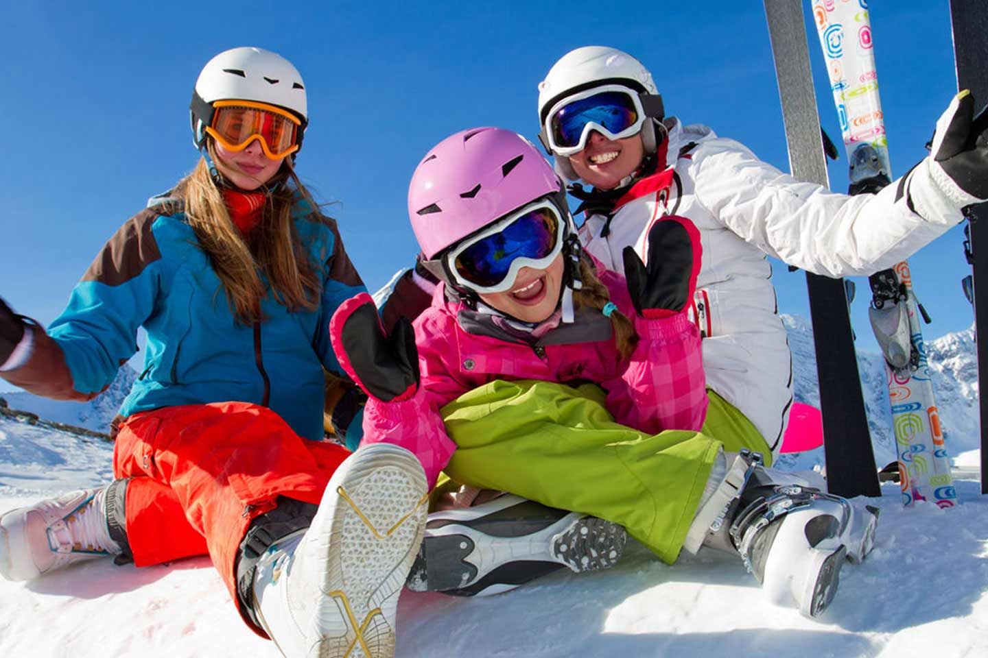 Drei Personen in bunter Skikleidung sitzen fröhlich im Schnee und posieren lachend vor strahlenden blauen Himmel.