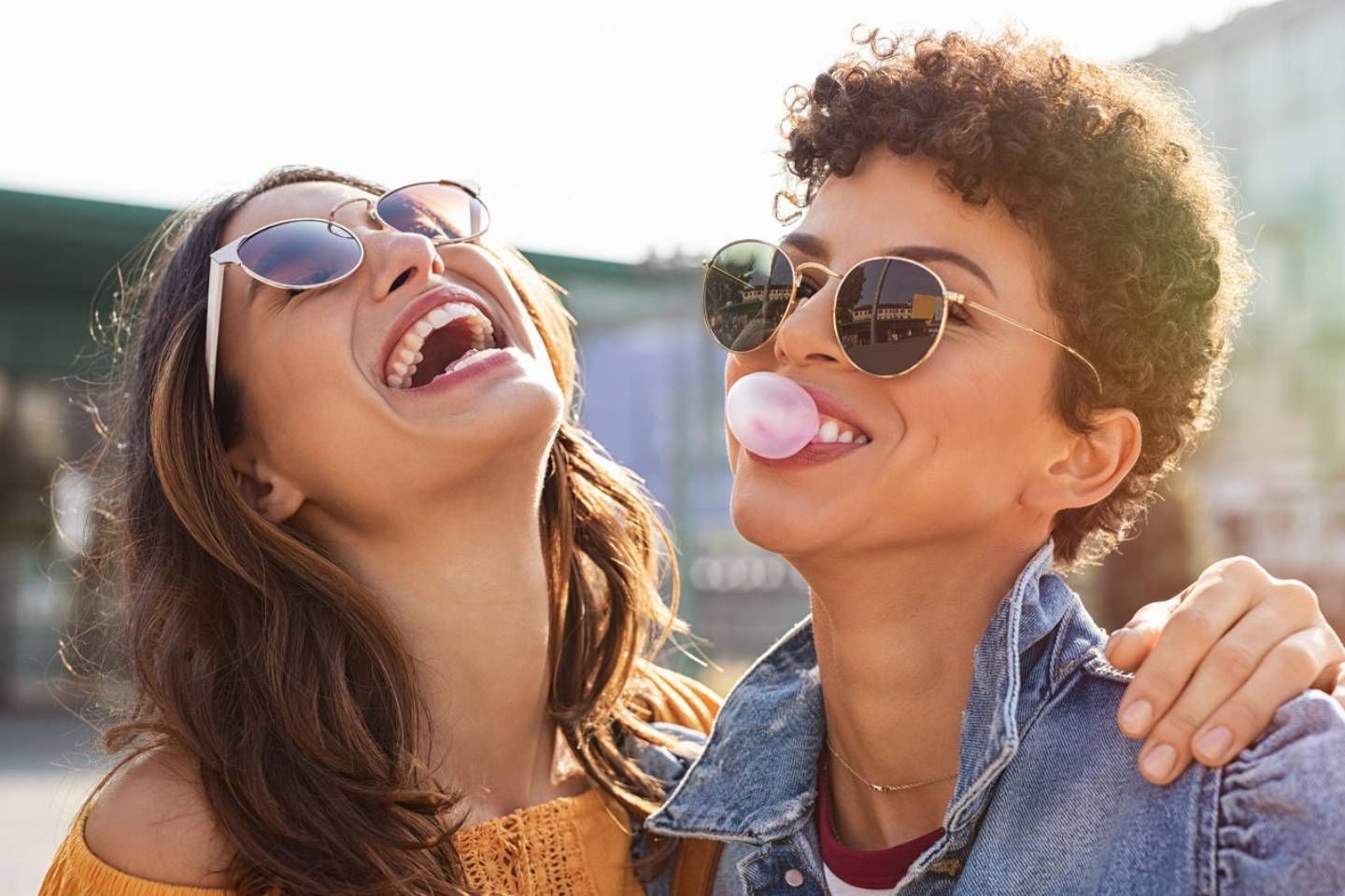 Zwei Frauen mit Sonnenbrillen lachen ausgelassen im Freien, während eine von ihnen Kaugummi zu einer Blase aufbläst.