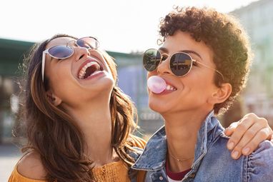 Zwei Frauen mit Sonnenbrillen lachen ausgelassen im Freien, während eine von ihnen Kaugummi zu einer Blase aufbläst.