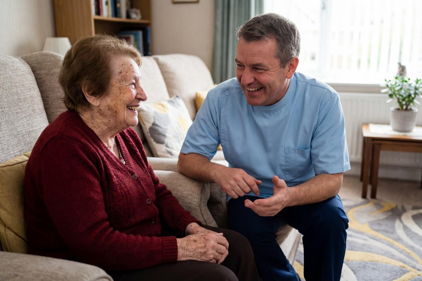 Ein Pflegeer und eine alte Dame sitzen lächelnd in ihrem Wohnzimmer.