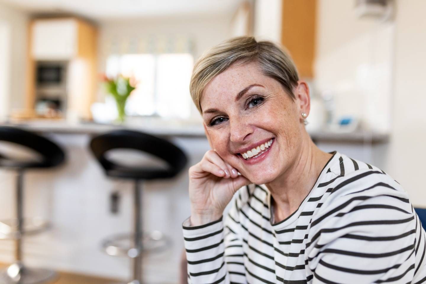 Eine lächelnde Frau mit kurzen, blonden Haaren und gestreiftem Shirt sitzt entspannt in einer modernen Küche. 
