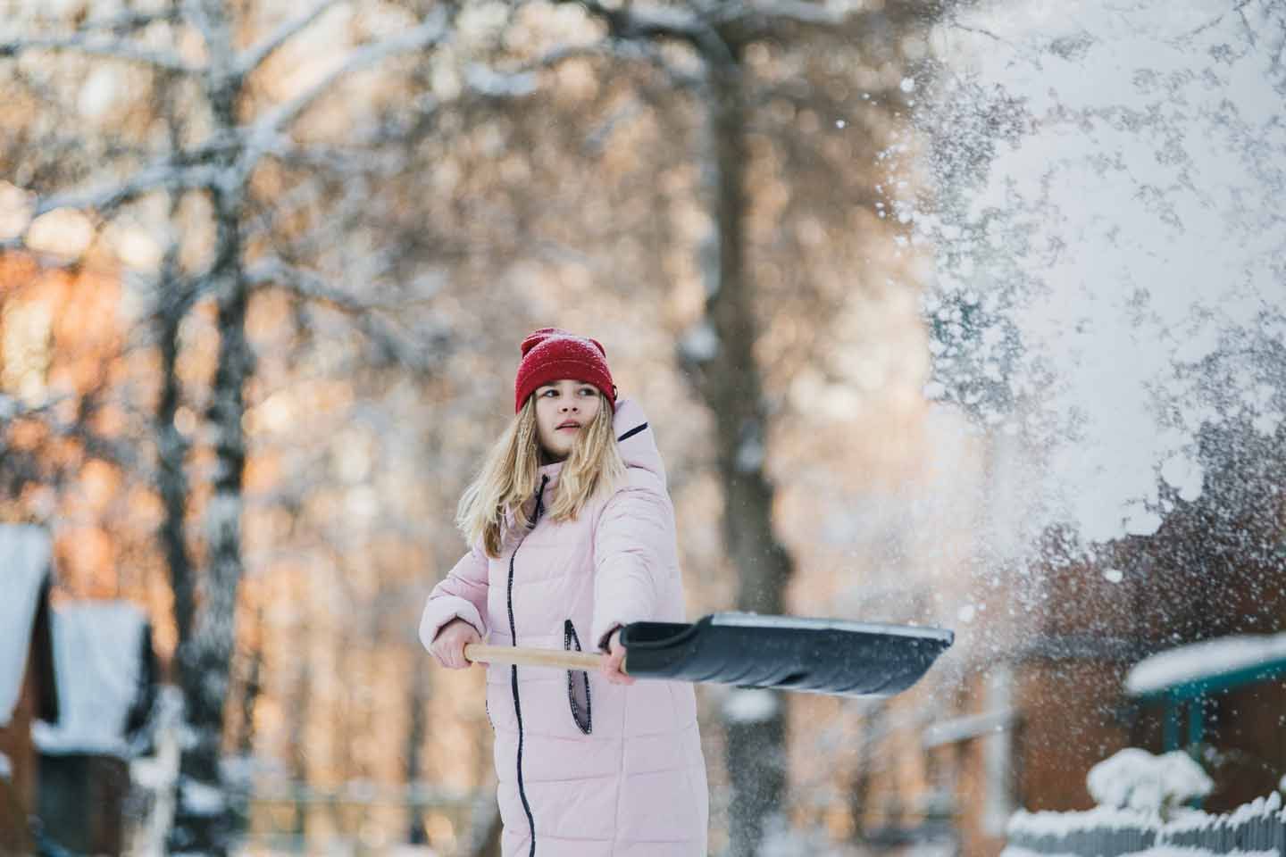 Eine Frau schippt im Winter Schnee. Sie trägt eine warme Jacke und Mütze.