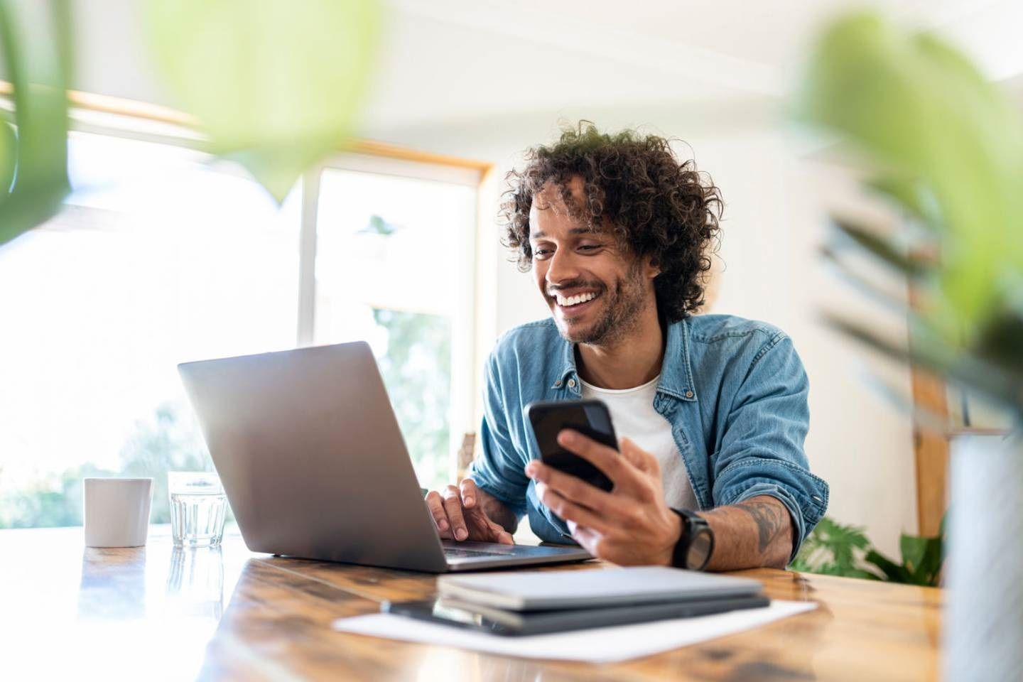 Ein junger Mann arbeitet zu Hause an einem Laptop und hält dabei ein Smartphone in der Hand.