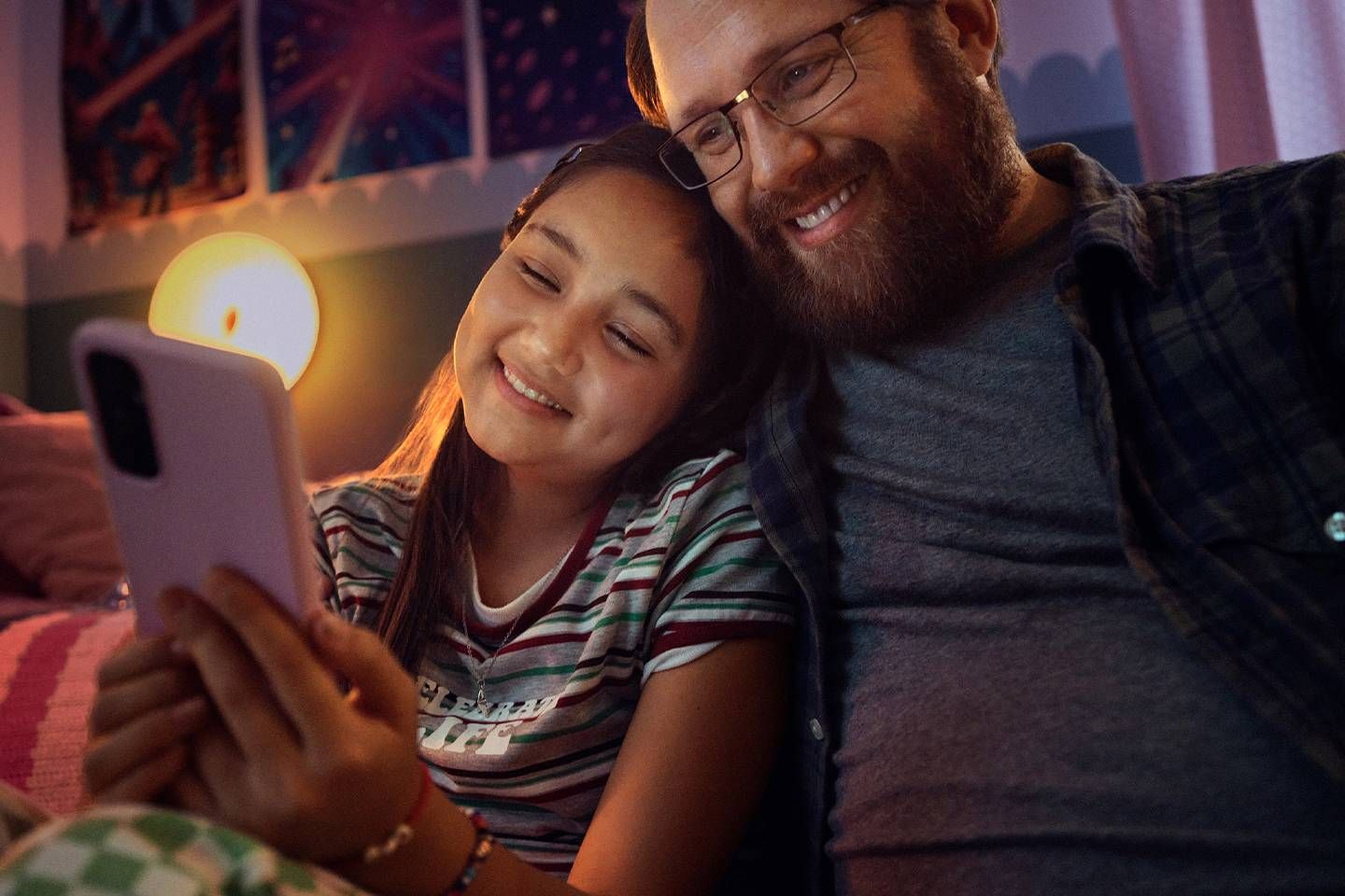 Mann und Mädchen sitzen eng beieinander auf dem Sofa und lächeln, während sie gemeinsam auf ein Smartphone schauen.