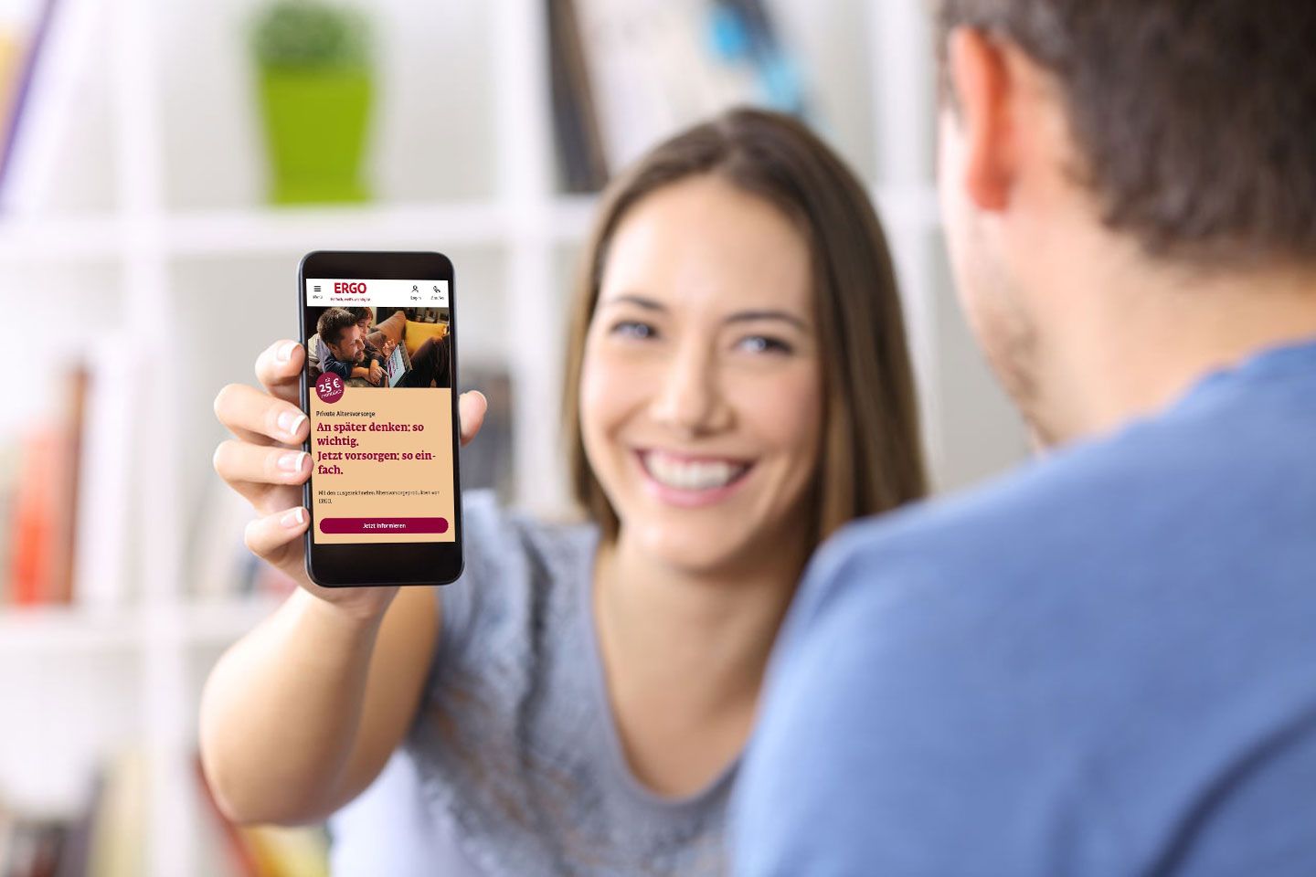 Junge Frau zeigt lächelnd ein Smartphone mit der ERGO-Website zur privaten Altersvorsorge, während sie mit einem Mann spricht.