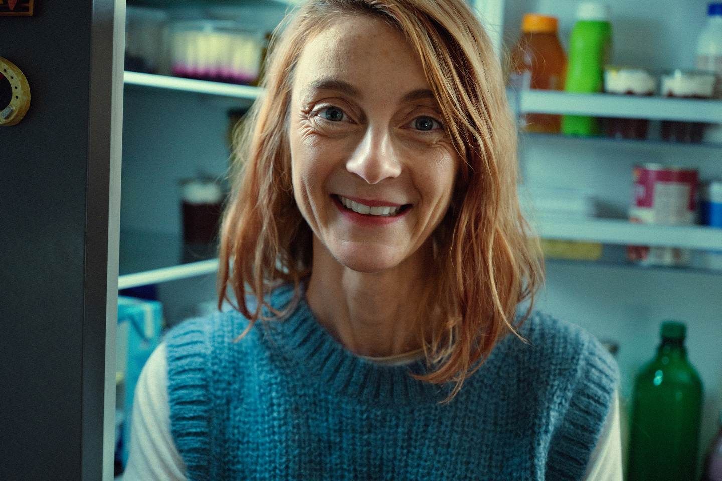 Lächelnde Frau steht vor einem geöffneten Kühlschrank mit verschiedenen Lebensmitteln im Hintergrund..