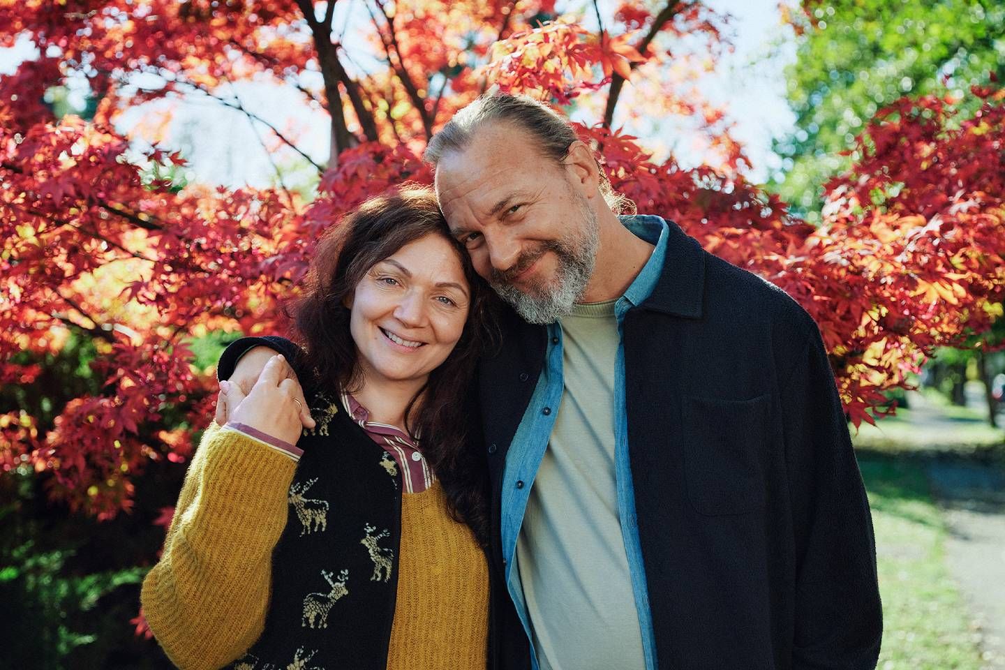 Lächelndes Paar steht eng umarmt im Freien vor herbstlich gefärbten Bäumen mit roten Blättern.