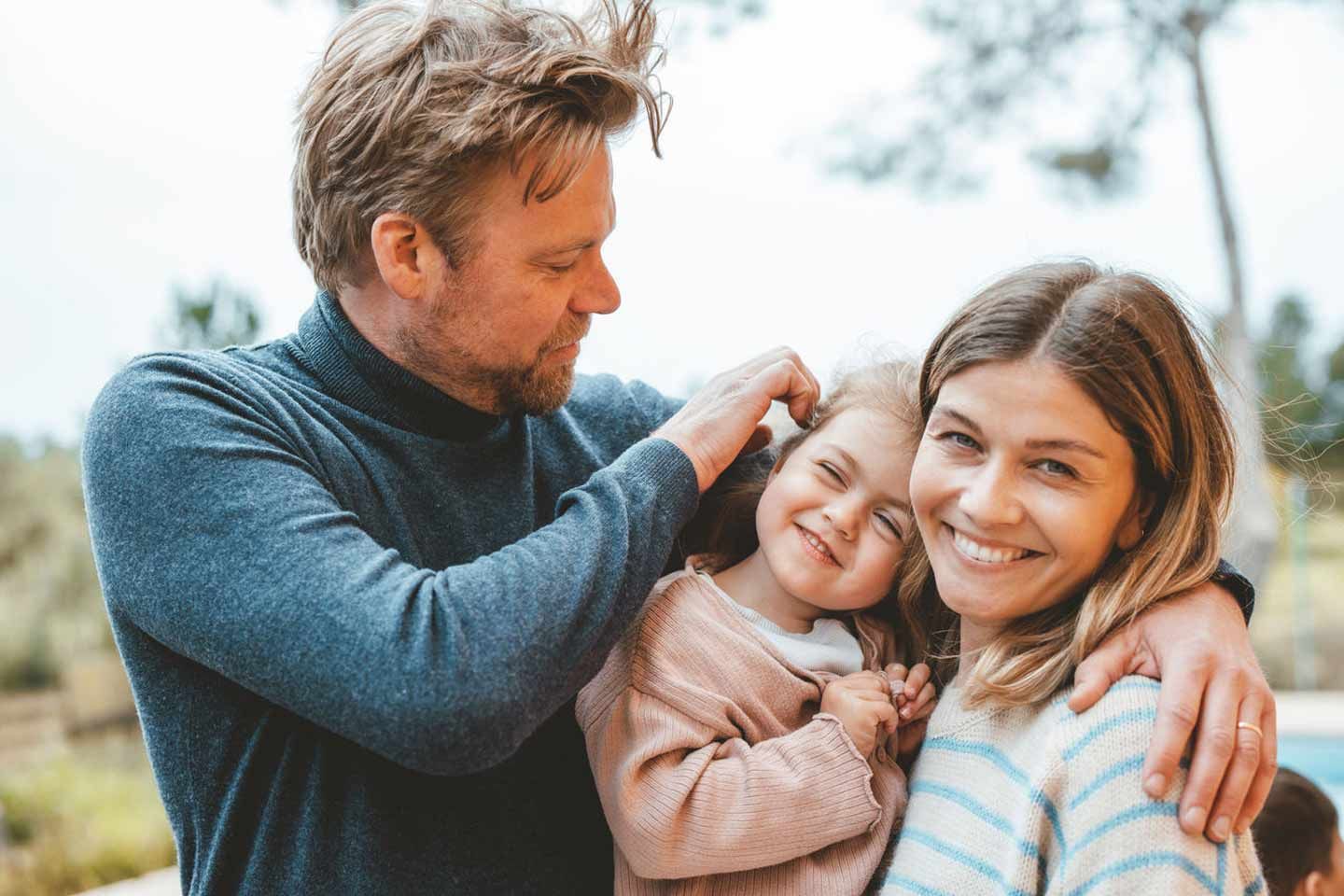 Junge Familie im Freien: Mutter und Vater umarmen liebevoll ihre lächelnde Tochter.