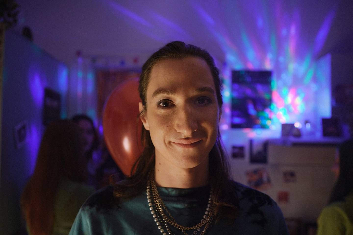 Junge Frau blickt lächelnd in die Kamera, während bunte Lichtreflexe und eine Partyatmosphäre den Raum im Hintergrund erhellen..