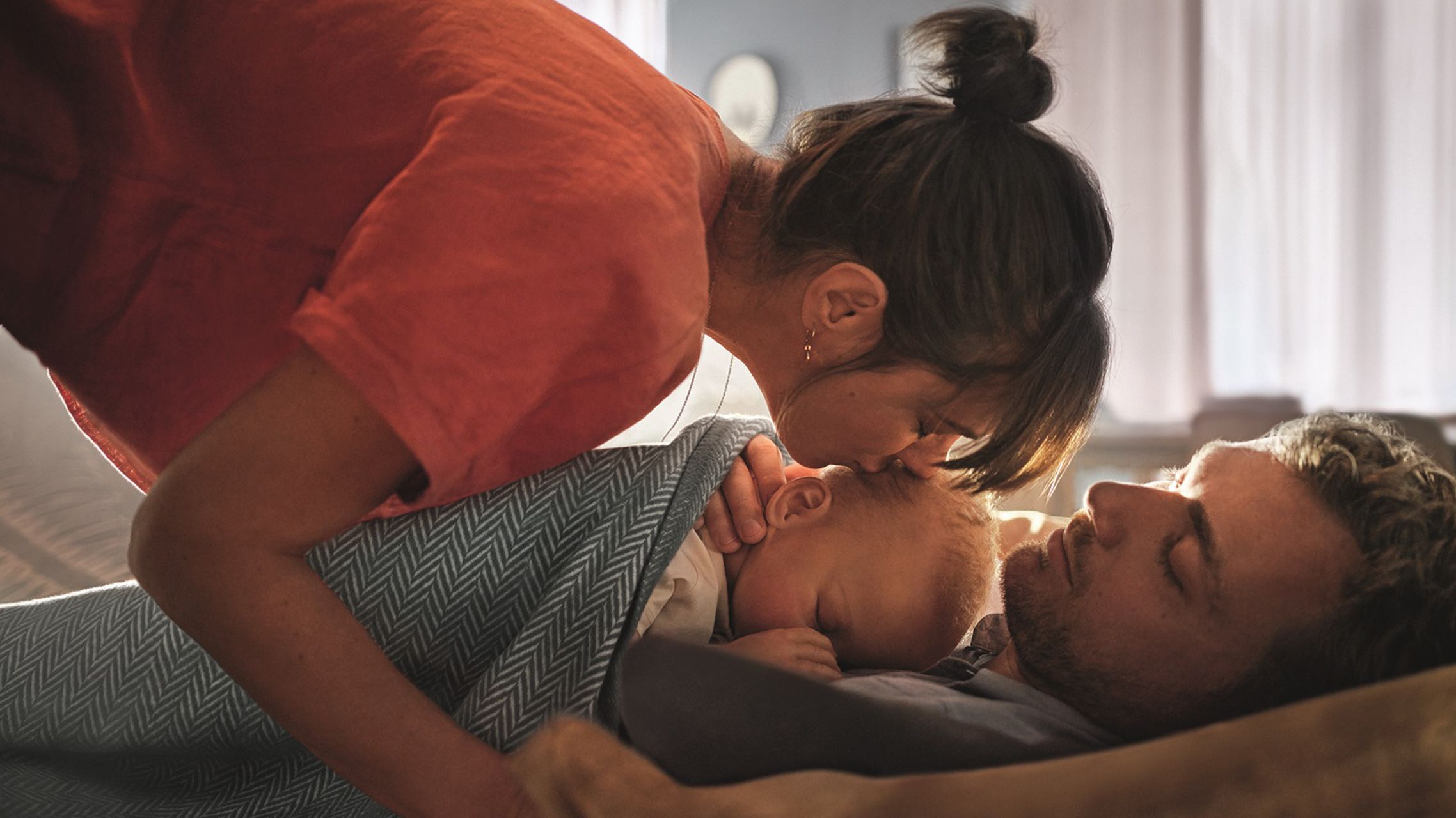 Frau gibt einem Baby einen Kuss, das auf dem Vater im Bett liegt.