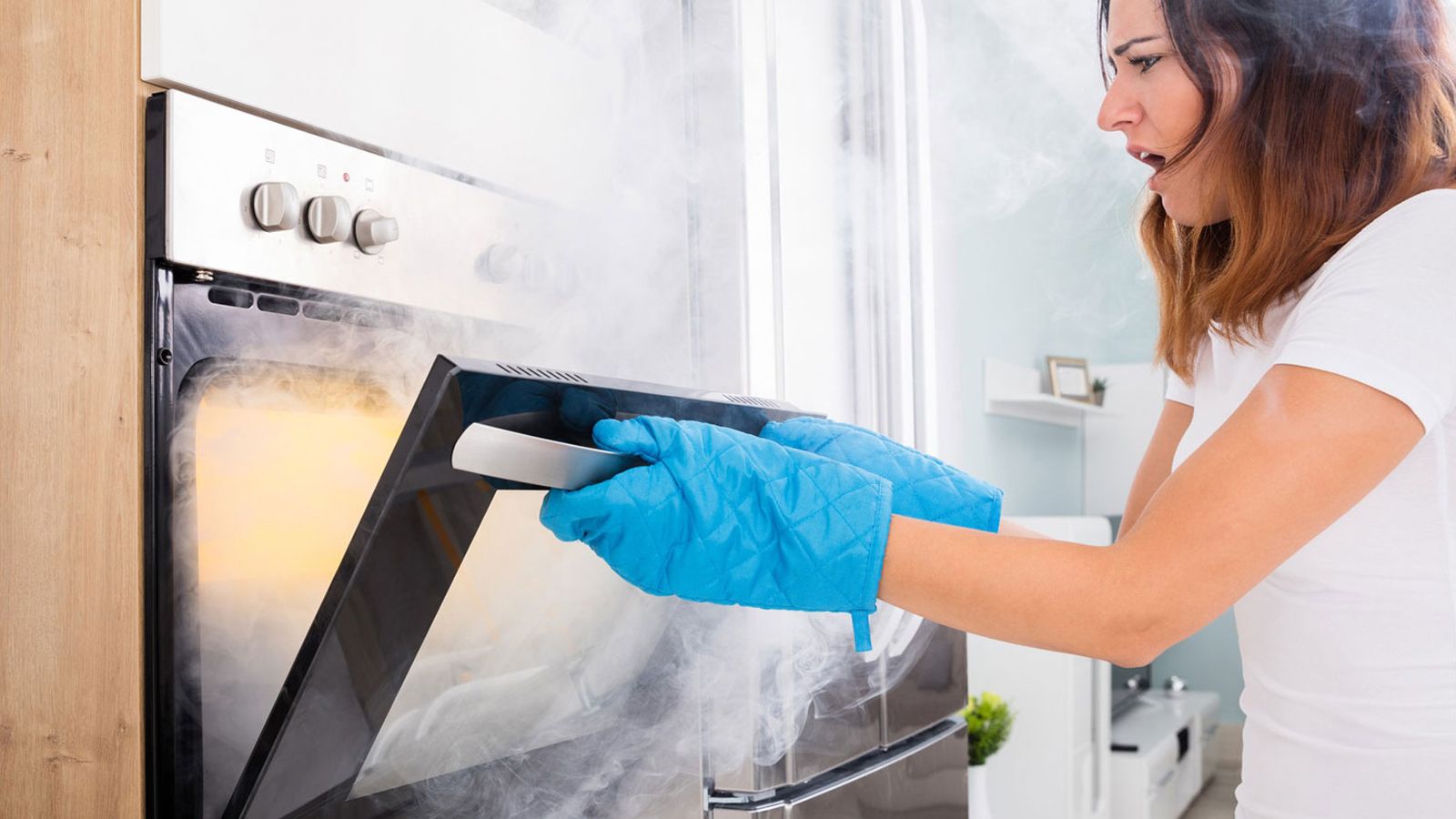 Eine Frau mit blauen Handschuhen öffnet einen Ofen, aus dem Rauch entweicht.