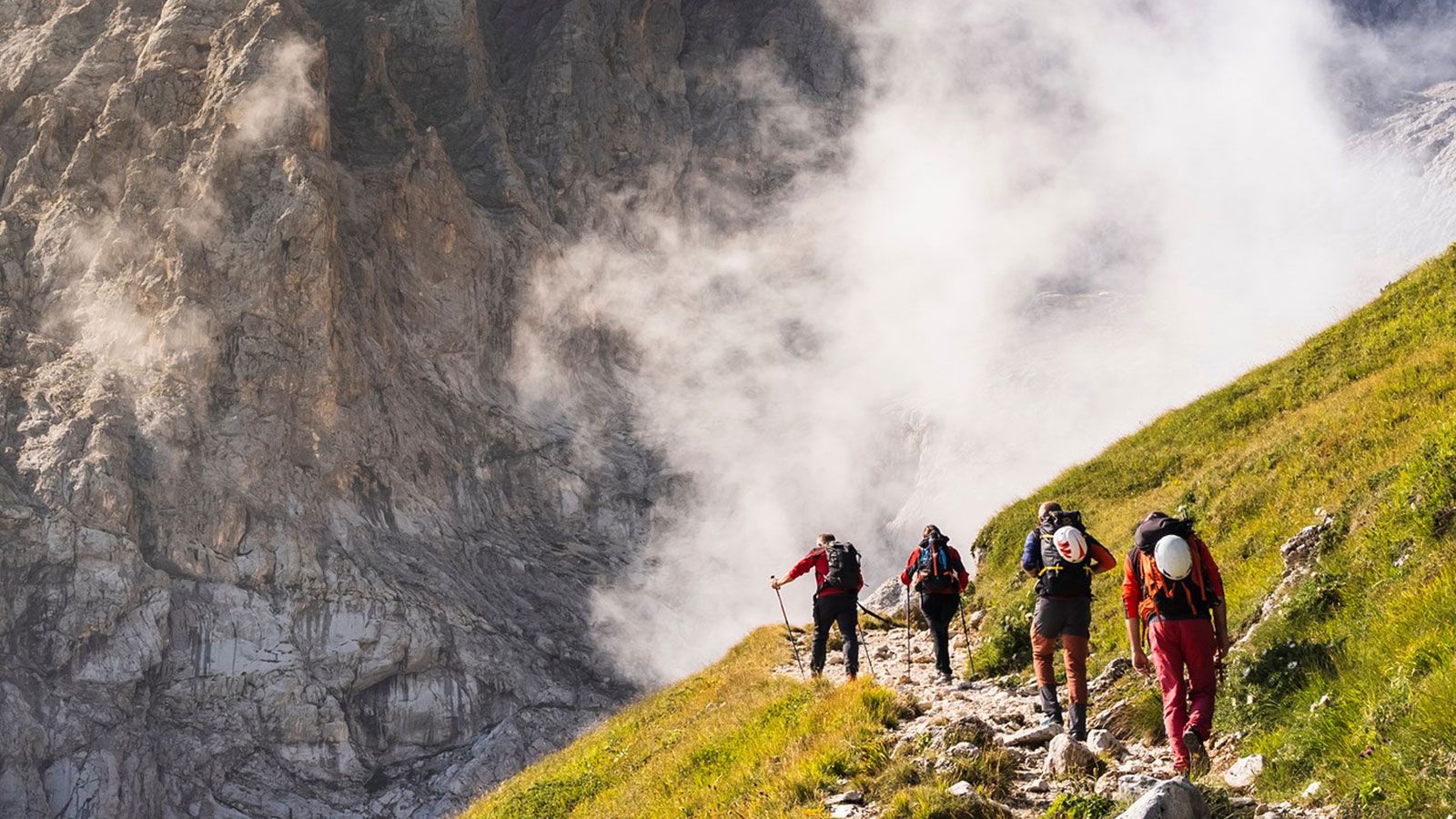 Wanderer auf einem Bergpfad, umgeben von grüner Natur und Bergen im Hintergrund.