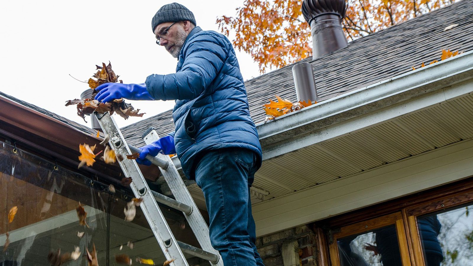 A man is cleaning the roof of leaves while standing on a ladder.