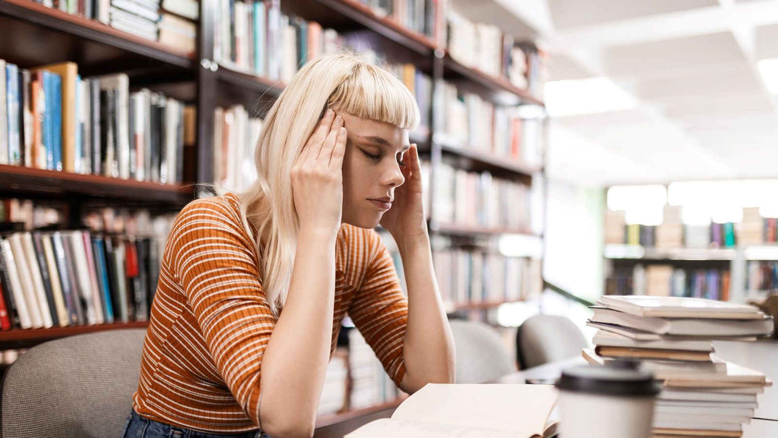 Eine Frau sitzt an einem Tisch, umgeben von Büchern, während sie sich an den Kopf fasst.