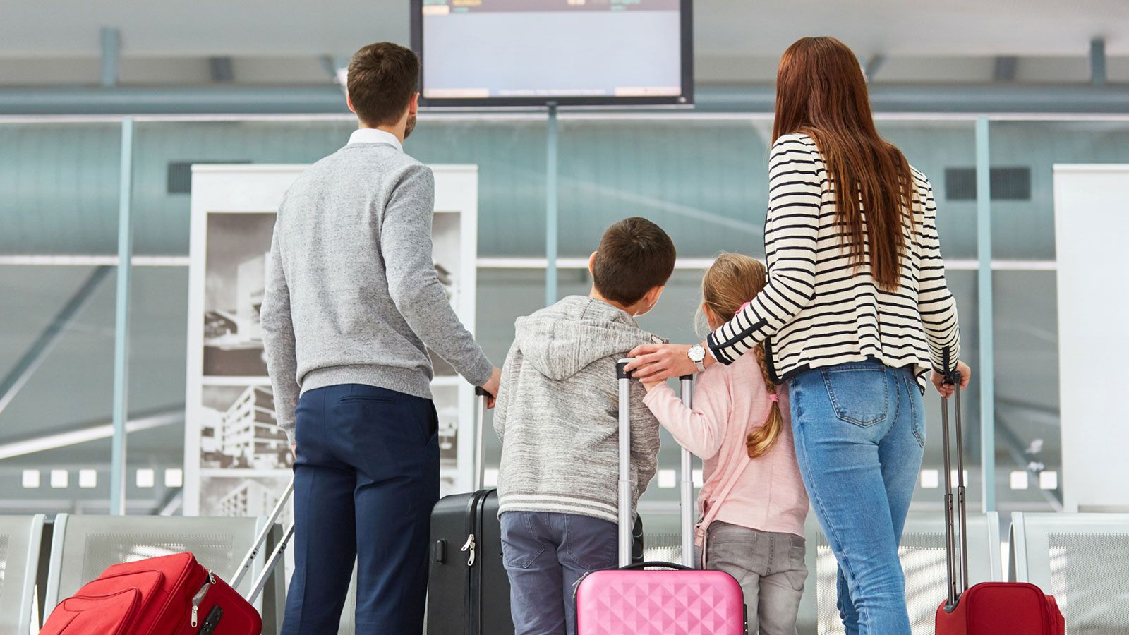 Familie mit zwei Kindern steht mit Rollkoffern vor einer Anzeigetafel in einem Flughafenbereich