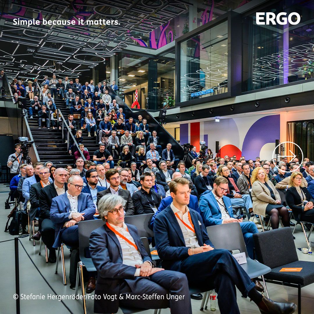 Many people are sitting as an audience on chairs and stairs in a room.