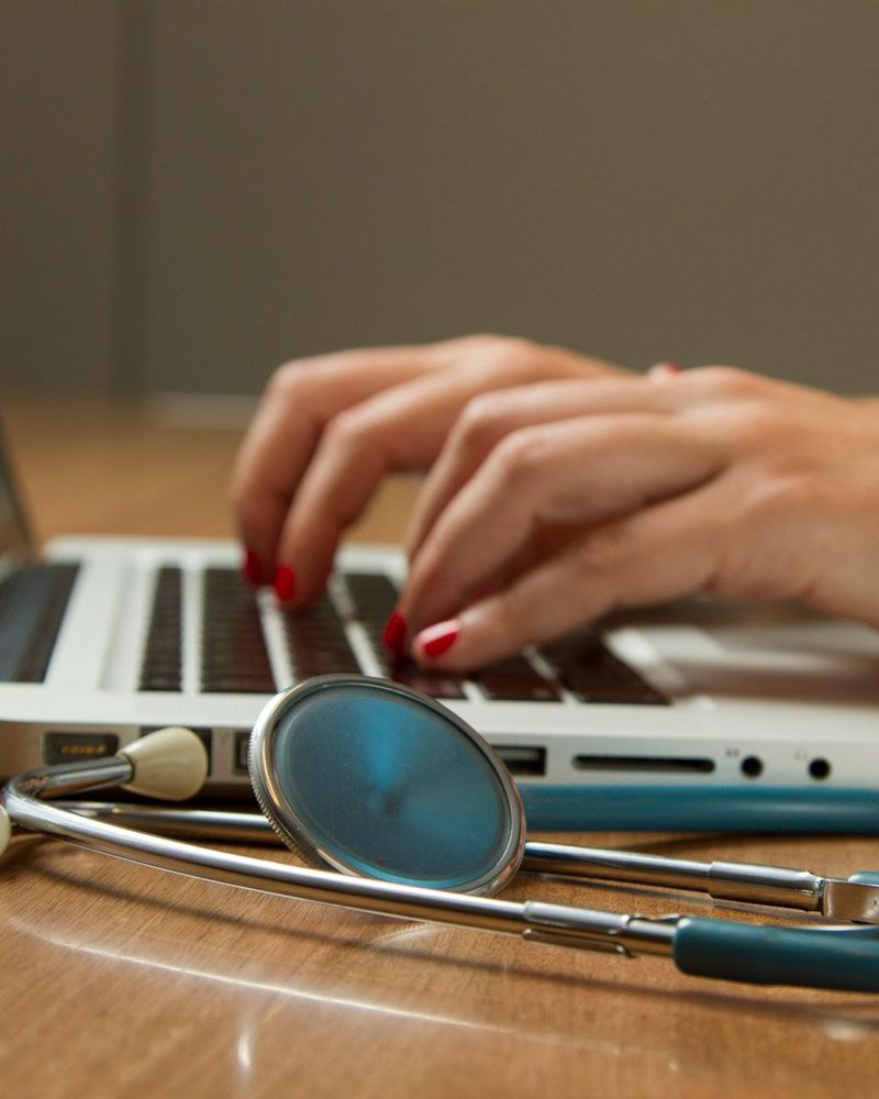 Two women’s hands are typing on a laptop. A stethoscope lies next to it.
