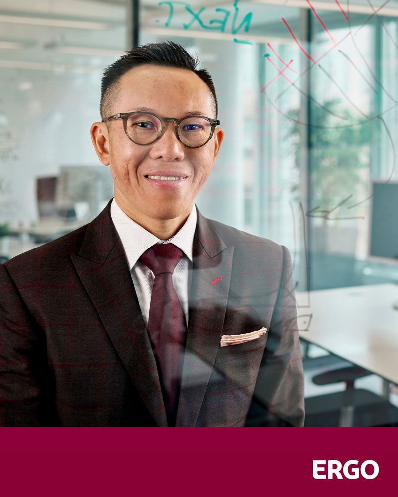 An Asian man in a suit is standing in the office, smiling at the camera.