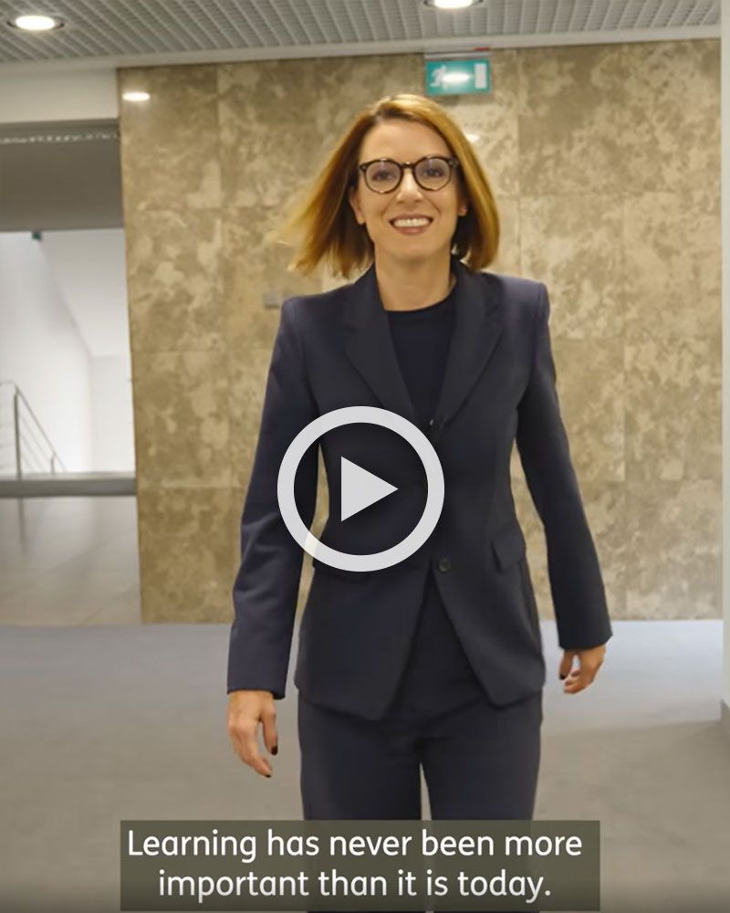Video still: A woman in a dark suit walks down a corridor 