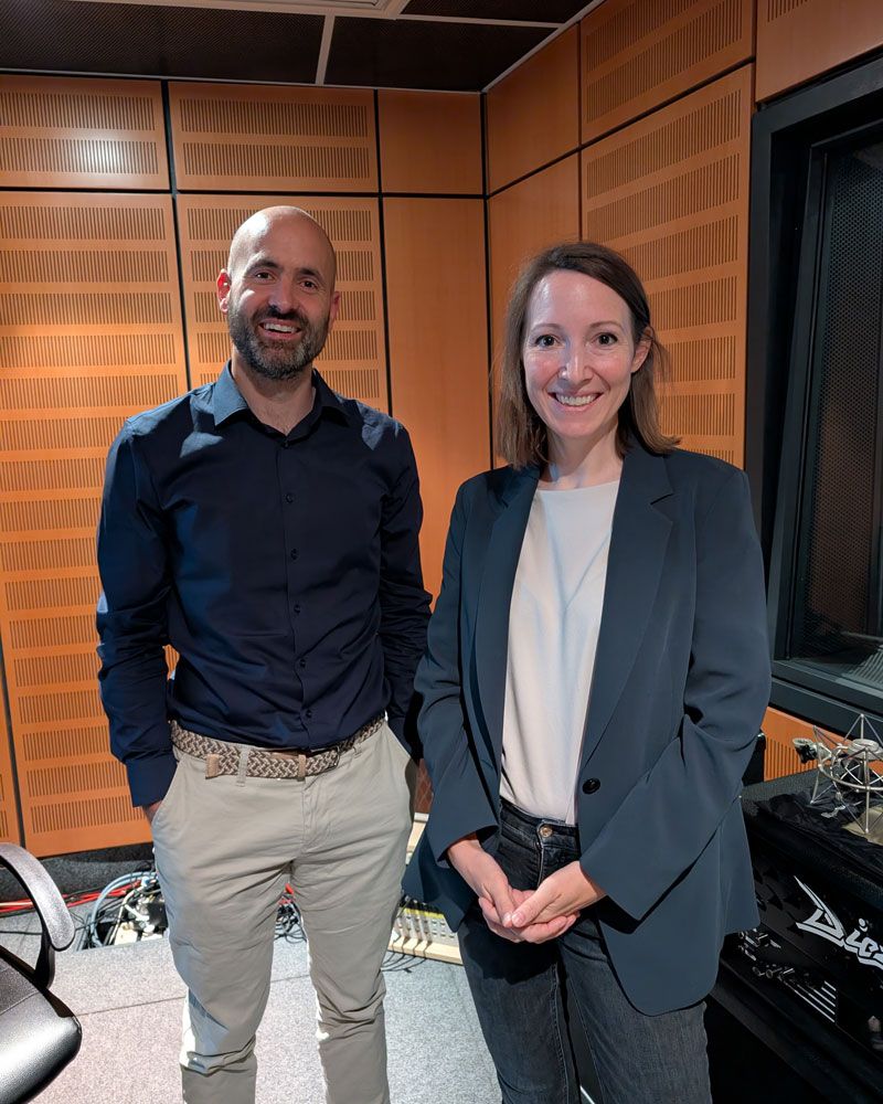 A man and a woman are standing in a recording studio, smiling at the camera.
