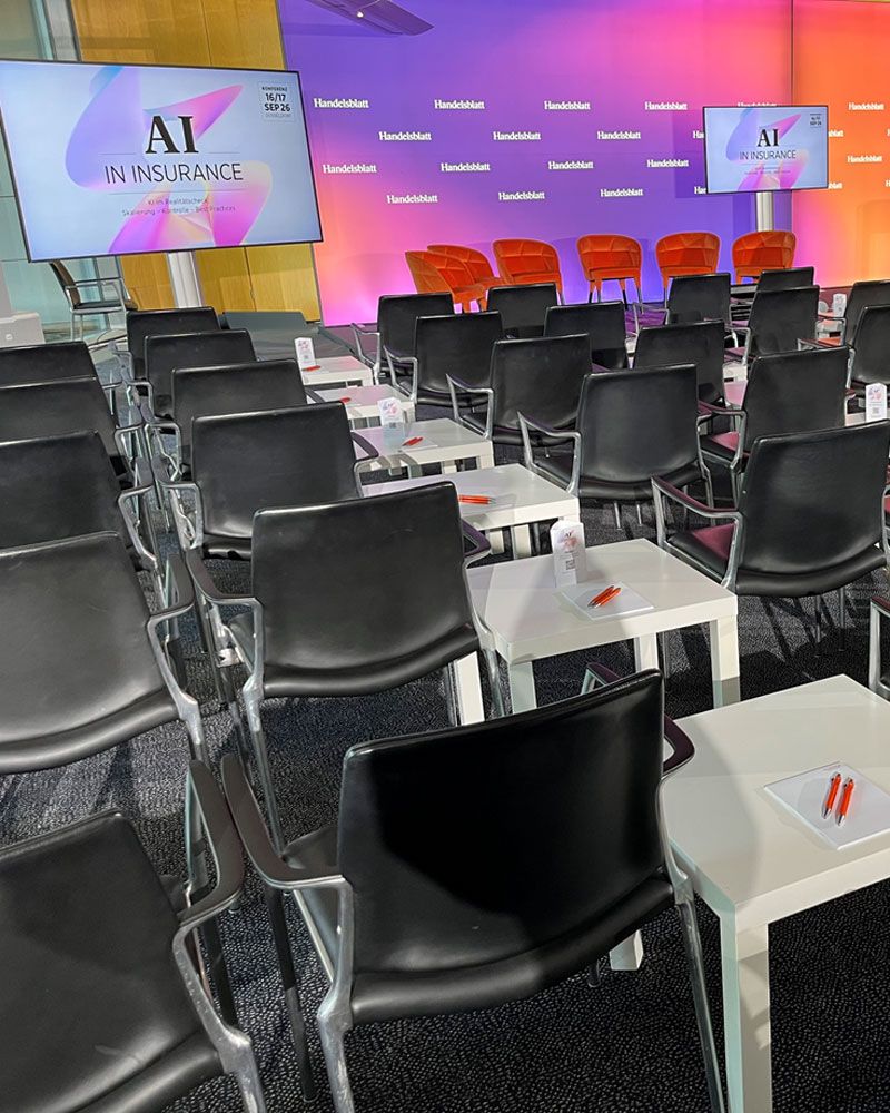 A conference room set up with rows of chairs, a colourful stage with red chairs and monitors